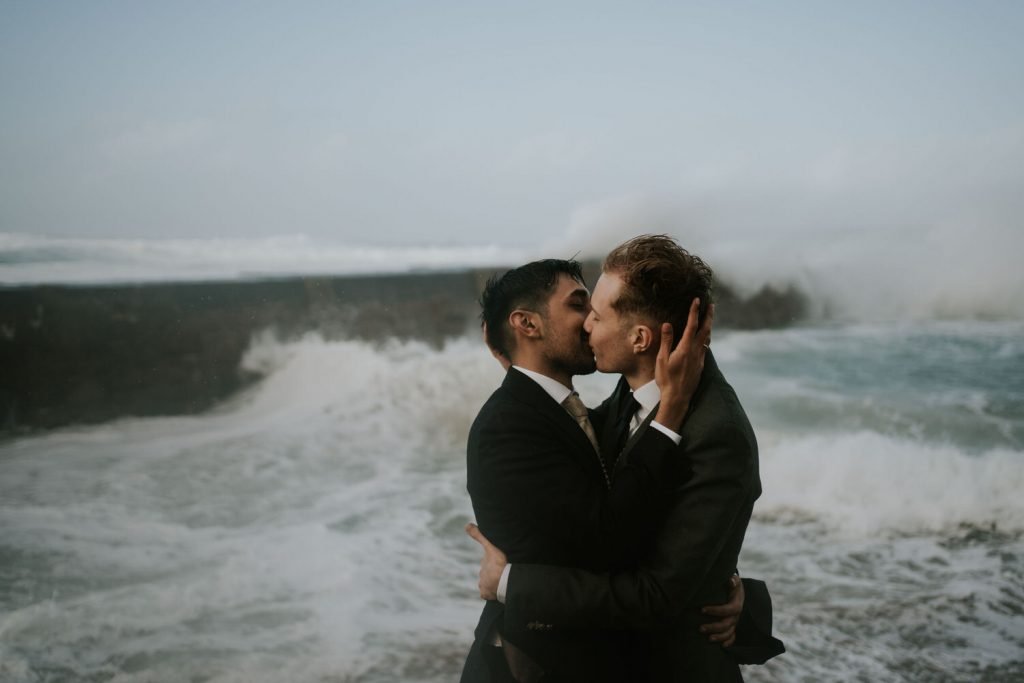 couple kiss during their micro wedding as a huge wave crashes behind them at Sennen Cove 