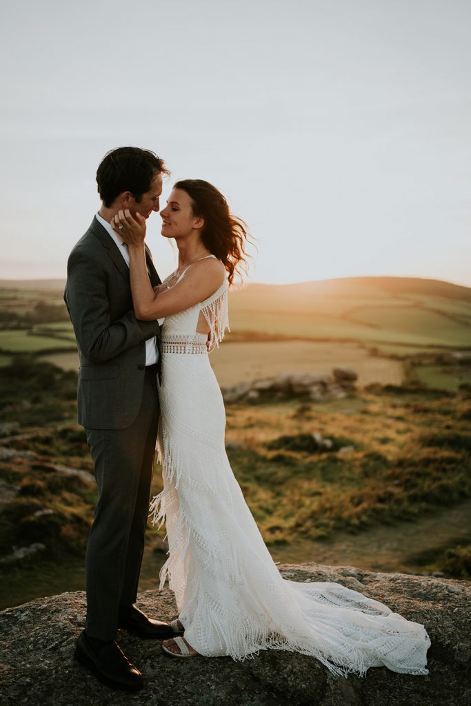 A bride and groom stand on a big rock at sunset as the wind gently blows the brides hair and she touches his face as they go in for a soft kiss 