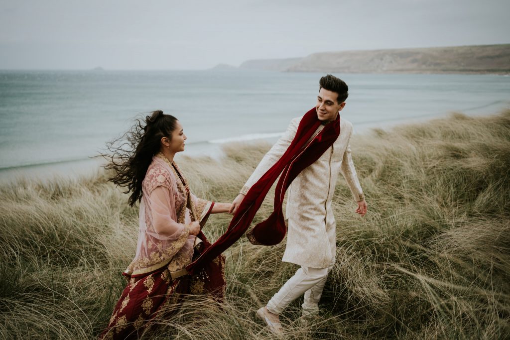 A bride and groom walk hand in hand through the sand dunes at Sennen Cove during their small wedding in Cornwall 
