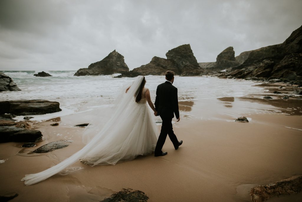 A couple walk hand in hand across a big dramatic beach in Cornwall as the brides dress drags behind in the wet sand 
