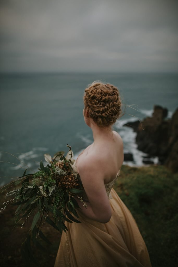 A bride holds dried and wild flowers in her hand, her hair braided and wearing a golden wedding dress 